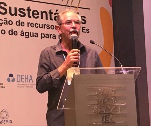 Um homem de cabelos grisalhos vestindo uma camisa cinza e segurando um microfone em um pódio, aparentemente falando para um público à frente dele.